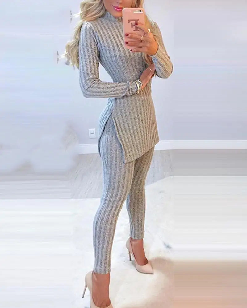 Posando elegantemente em frente a um espelho, uma pessoa exibe o conjunto para inverno de Selluve, com um vestido suéter cinza canelado com leggings combinando e saltos altos nude. O cabelo cai em ondas soltas enquanto ela segura um telefone rosa contra o fundo interior suavemente desfocado.