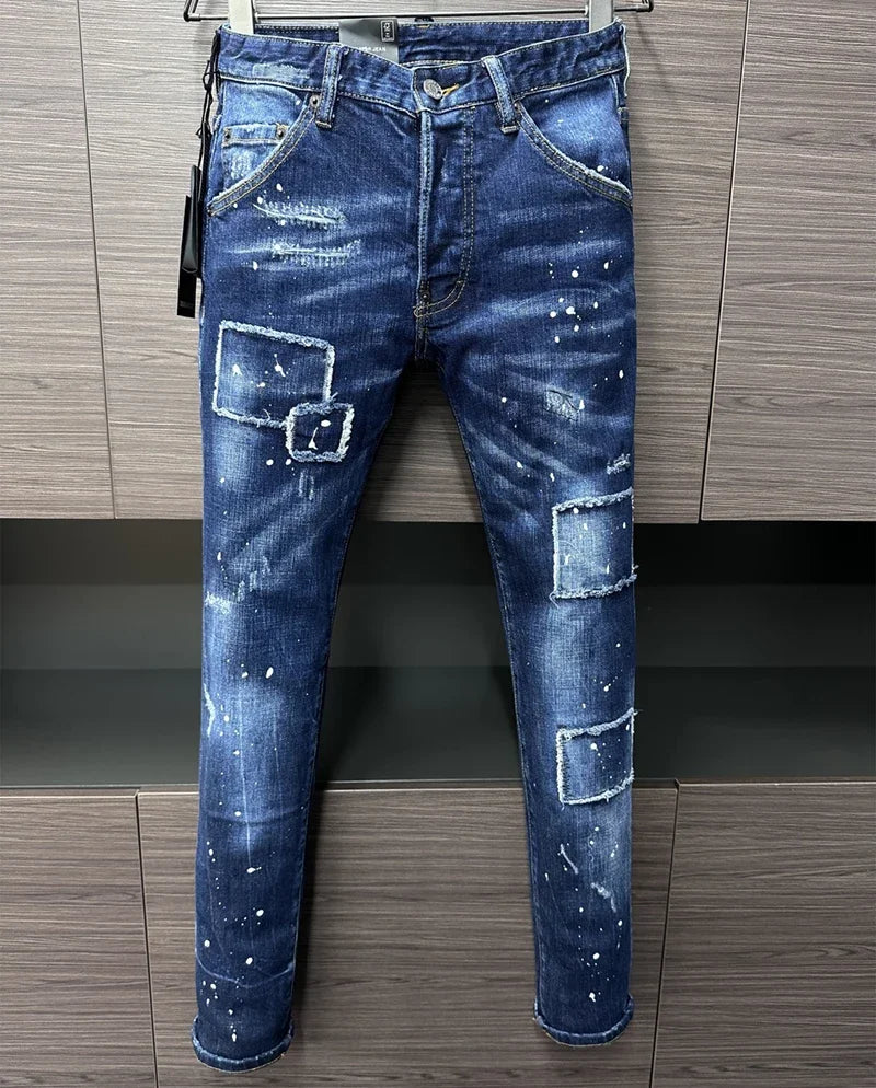 A calça jeans masculina BRAND DSQ2 da Selluve apresenta um corte justo com respingos de tinta e vários remendos retangulares, sobre um fundo de painel de madeira cinza.