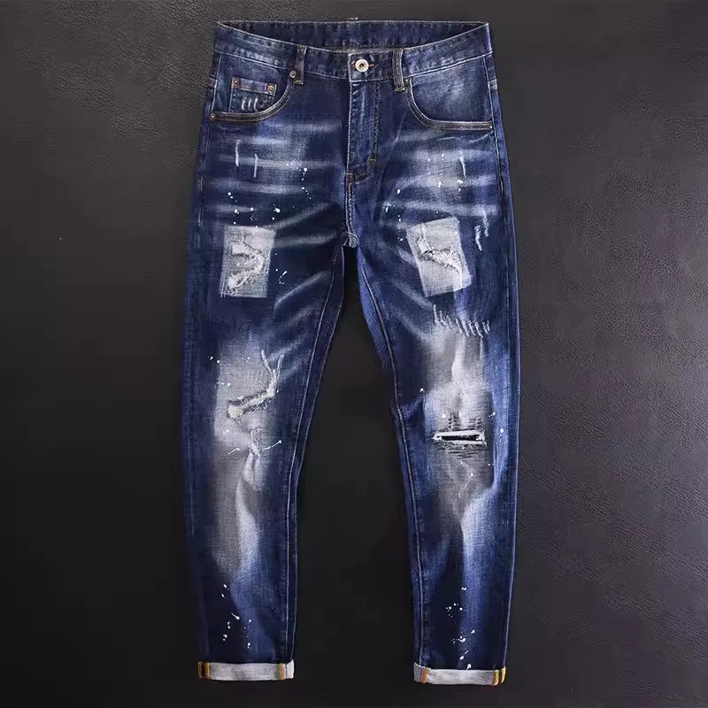 Jeans masculinos vintage elásticos da Selluves Street Fashion em azul desgastado com punhos enrolados, respingos de tinta e remendos são colocados sobre um fundo escuro. Esses jeans incorporam o estilo hip-hop com um ajuste fino vintage, ideal para o criador de tendências urbanas.