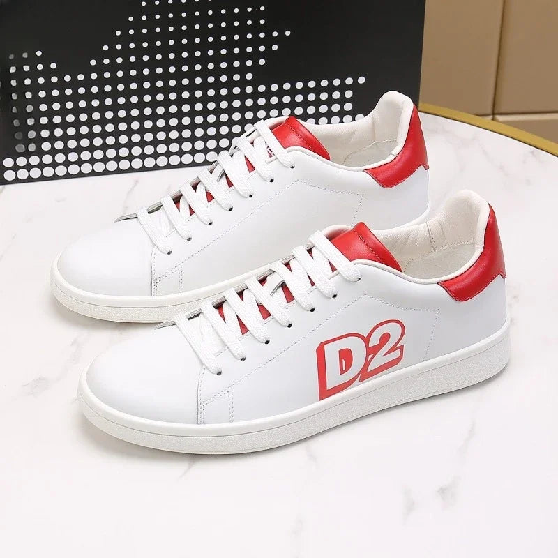 Os D2 Sapatos Masculinos Luxo da Selluve são tênis brancos com detalhes em vermelho no calcanhar e na língua, exibindo D2 em vermelho forte na lateral. Feitos de couro genuíno, eles exemplificam elegância e conforto e repousam sobre uma superfície de mármore branco contra um fundo preto perfurado.