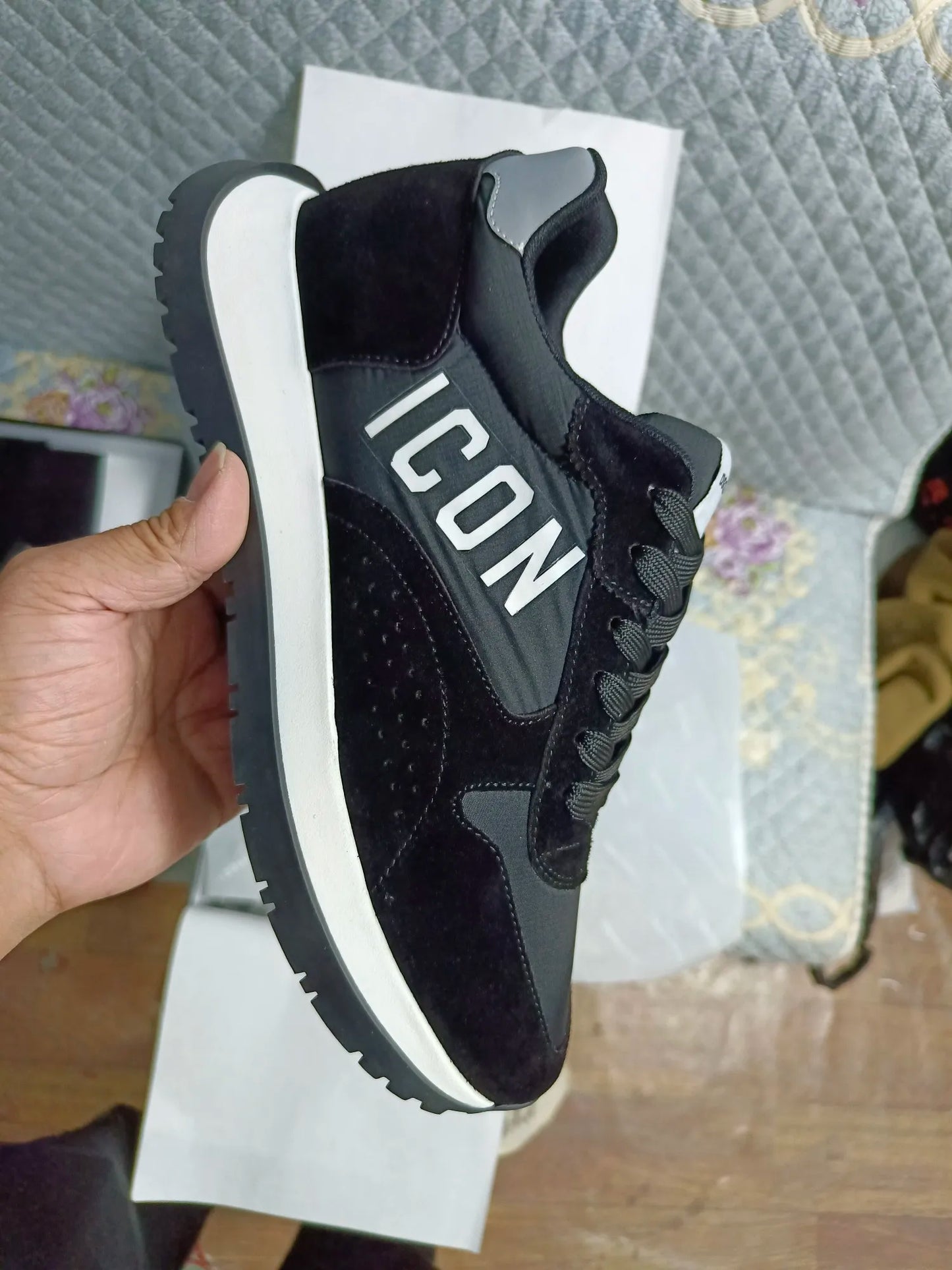 Uma mão segura um tênis masculino ICON da Selluve, com "ICON" impresso na lateral. Este sapato masculino preto e branco incorpora conforto e elegância com seu design texturizado sobre uma superfície acolchoada.