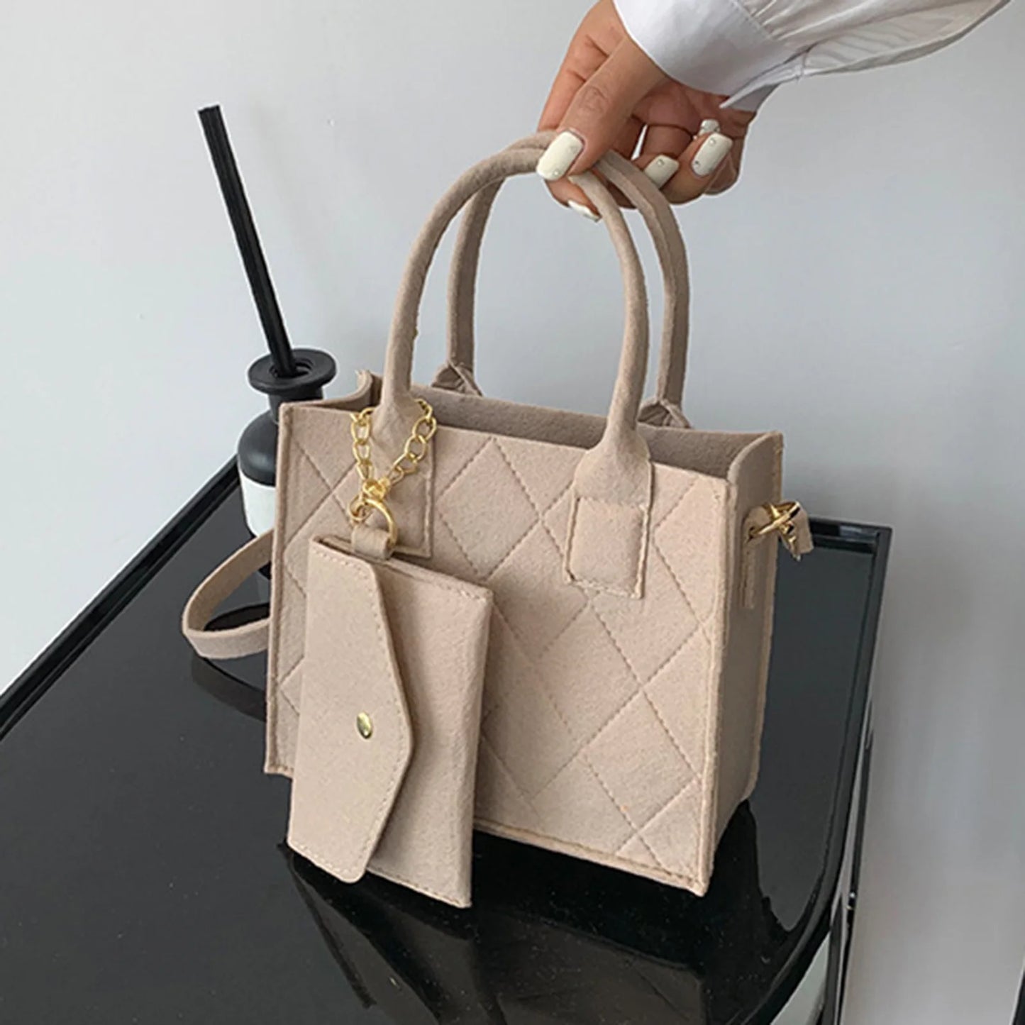 Uma mão exibe uma "Bolsa de feltro feminina" bege acolchoada da Selluve com uma corrente dourada e carteira combinando em uma superfície preta. Um copo de palha preto é parcialmente visível no fundo. A pessoa, usando esmalte branco e uma blusa branca de manga comprida, completa este conjunto chique.