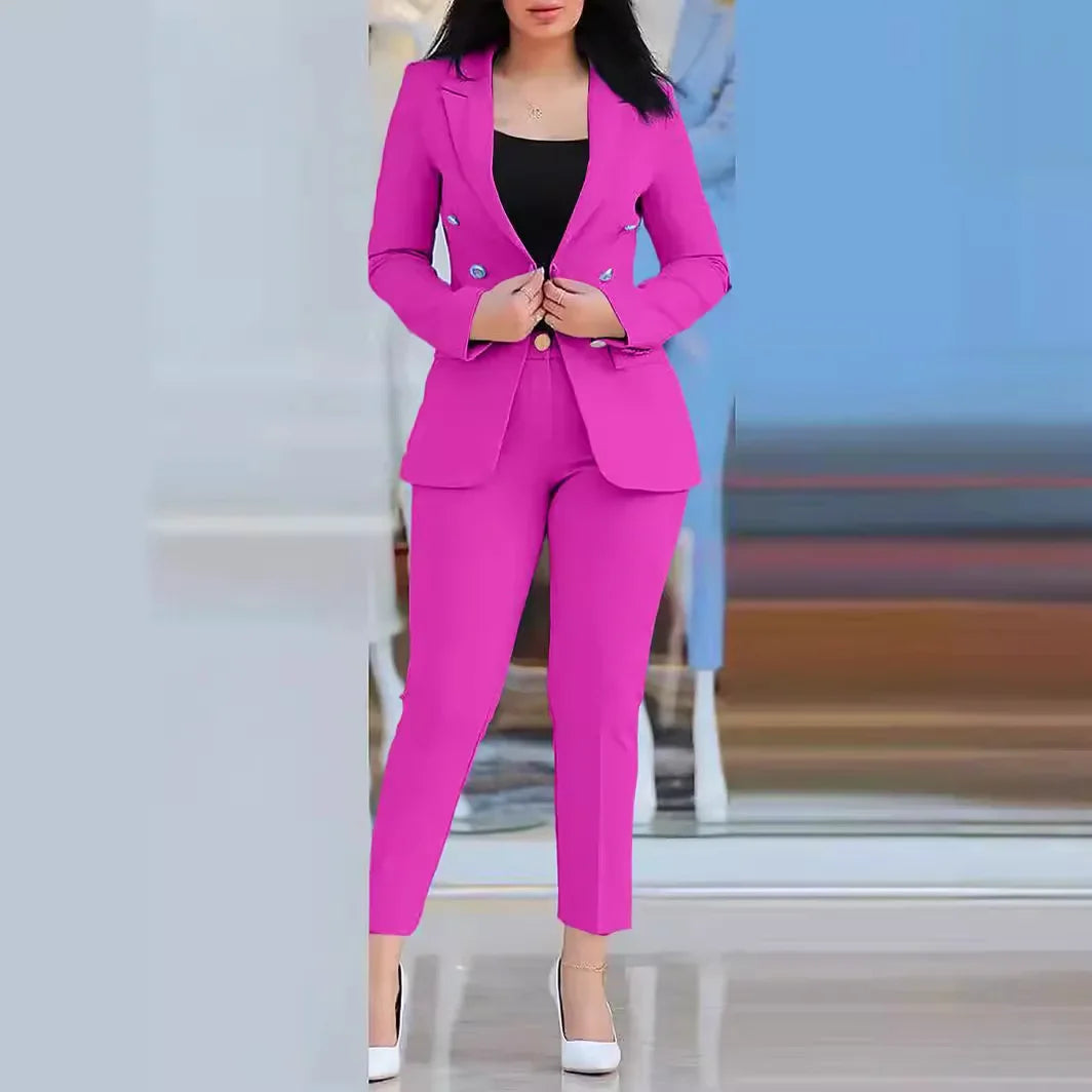 Contra um fundo moderno e desfocado, alguém veste casualmente um terninho rosa vibrante, com calças de cintura alta e saltos brancos, da coleção "Roupas de Escritório para Mulheres", da Selluve. O blazer justo é usado sobre uma blusa preta, com as mãos ajustando as mangas compridas.