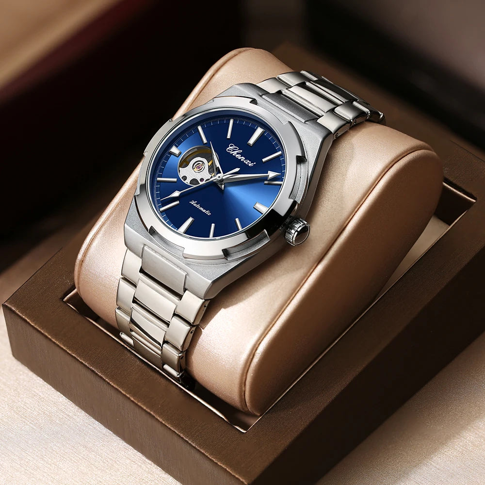 Um relógio de pulso mecânico Selluve prateado com design de coração aberto e mostrador azul assenta elegantemente sobre um suporte almofadado castanho, exibindo o seu mecanismo de estilo e durabilidade com uma bracelete metálica.