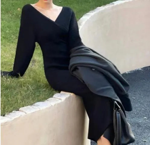 Uma pessoa usando um vestido preto High End Matching da Selluve com decote em V está sentada em um muro de pedra curvado, segurando um casaco preto e uma bolsa. A grama atrás do muro e um caminho pavimentado de um lado sugerem um jardim ao ar livre ou um cenário de parque.