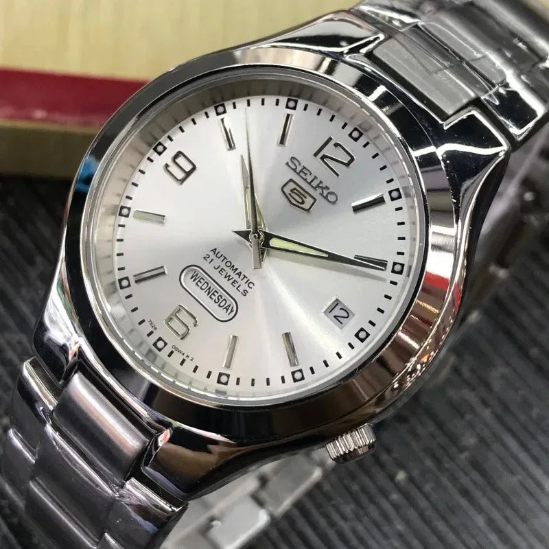 O Selluve Seiko Luminous Calendar Automatic apresenta um design sofisticado com uma faixa metálica prateada e aro polido. Seu mostrador branco redondo tem marcadores de hora prateados, ponteiros e funções de dia-data às 3 horas, simbolizando a elegância de um relógio automático.