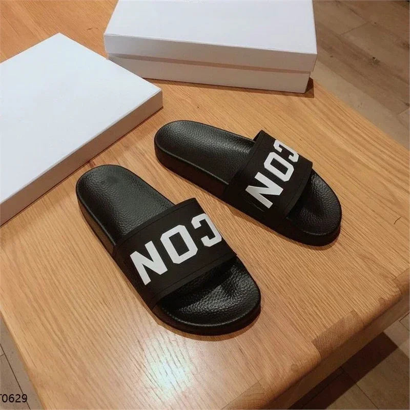 Chinelos masculinos pretos Selluve ICON Letter com ICON nas tiras ficam sobre uma mesa de madeira, feitos de EVA premium para luxo. No fundo, duas caixas brancas complementam este elegante chinelo masculino projetado para conforto prolongado.