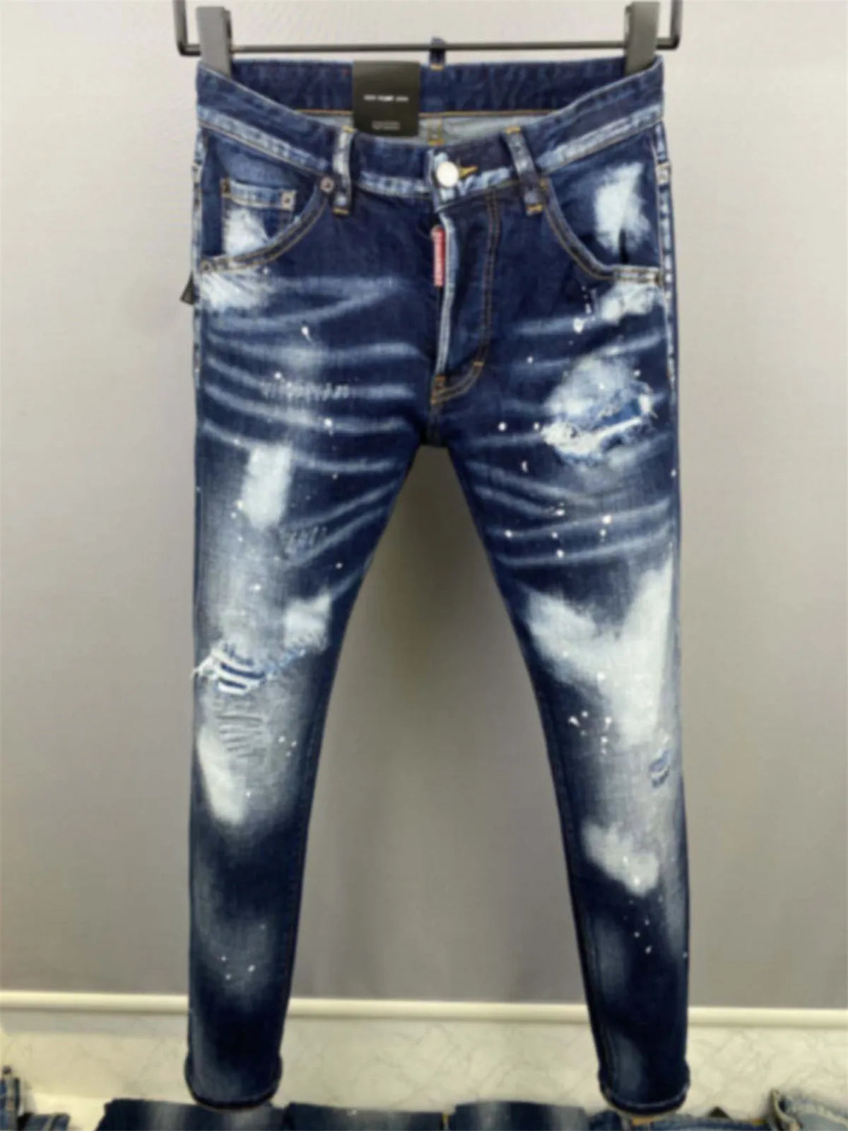 Um par de jeans Novo Masculino Azul Claro D2 da Selluve está pendurado em um fundo cinza, exibindo jeans azul desgastado com lavagem desbotada, remendos desfiados, manchas brancas para um estilo único, bolsos padrão e braguilha com botões.